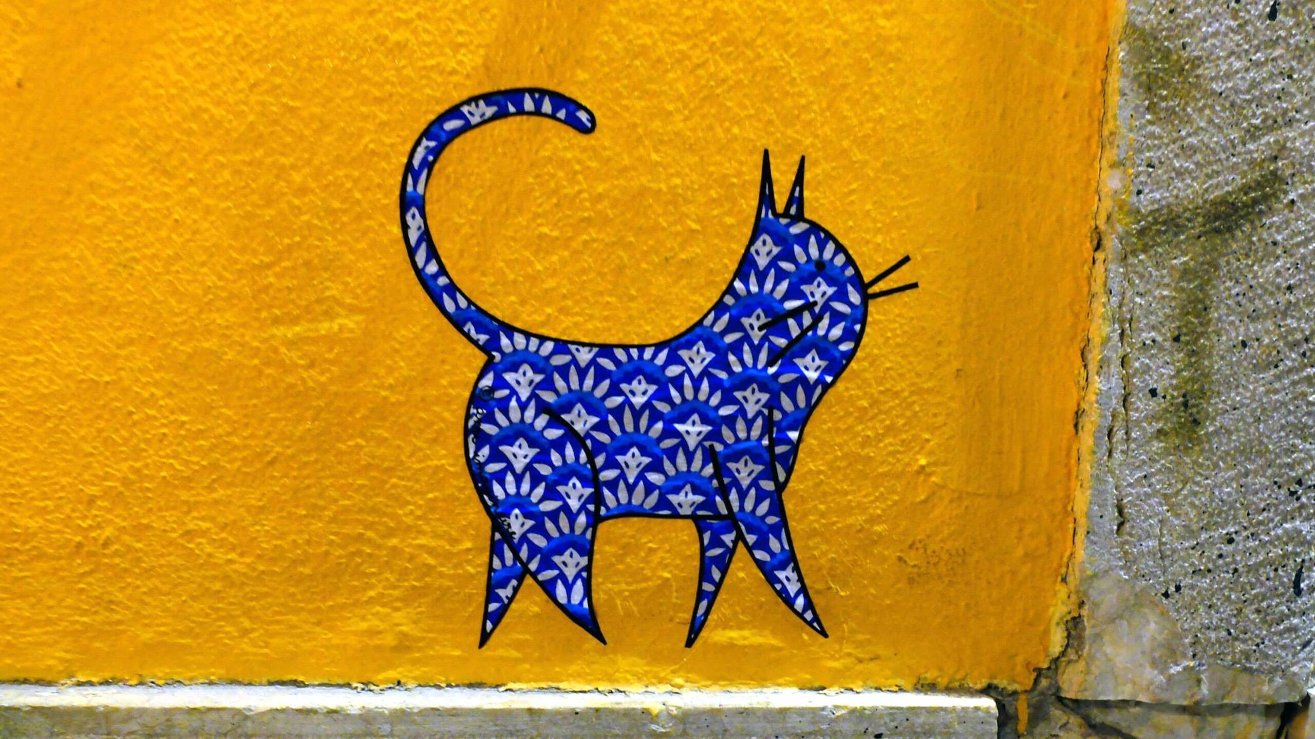 Oeuvre murale représentant un chat bleu sur un mur jaune. Oeuvre réalisée par la street artiste Ami Imaginaire.
