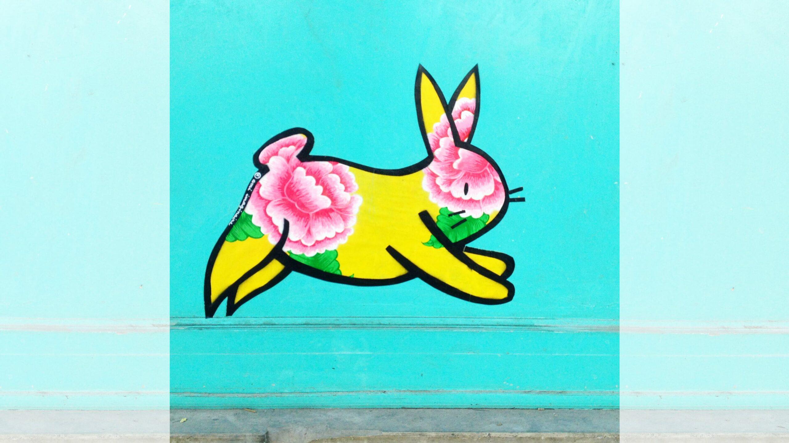Oeuvre de la street artiste parienne Ami imaginaire représentant un lapin jaune décoré de pivoines sur un mur bleu clair.
